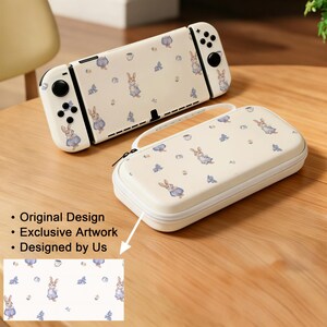 Könnte beinhalten: Cremefarbene Nintendo Switch Konsole und Tragetasche, beide mit einem sich wiederholenden Muster aus Aquarellhasen und blauen Akzenten verziert. Die Tasche hat einen Reißverschluss und einen Griff. Der Text im Bild lautet: Original Design, Exclusive Artwork, Designed by Us.