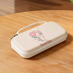 May include: A cream-colored rectangular case with a white zipper and handle. The case features a cartoon bunny holding a pink tulip, with the words "Good luck" printed below. The design includes stars and a purple bow.