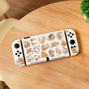 May include: A white Nintendo Switch console with a playful cat-themed design. The console features a cream-colored body and controllers adorned with various cartoon cat illustrations in different poses. The cats are in shades of orange, gray, and white.