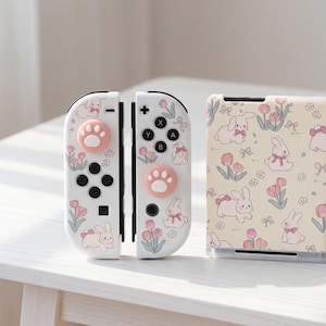 Puede incluir: Mandos Joy-Con blancos para Nintendo Switch con un diseño de conejitos y flores. Los mandos tienen botones rosas con huellas de patas y una funda a juego. El diseño incluye conejos, flores y lazos, creando una estética linda y juguetona.