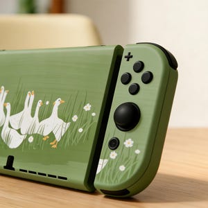 Könnte beinhalten: Eine grüne Nintendo Switch-Hülle mit einem Design aus weißen Gänsen in einem Feld mit Gras und weißen Blumen. Die rechte Seite der Hülle hat schwarze Knöpfe und einen schwarzen Joystick. Die Hülle befindet sich auf einer Holzoberfläche.