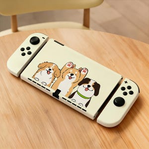 May include: A cream-colored handheld gaming console featuring a decorative skin with a cartoon illustration of three dogs. The console includes two detachable side controllers with black joysticks and buttons, all displaying a clean, smooth finish.