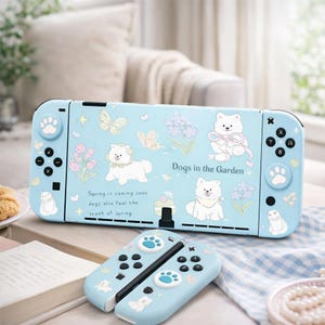 Puede incluir: Una consola y mandos Nintendo Switch de color azul claro con un diseño temático de perros. La consola presenta ilustraciones de perros blancos, mariposas y flores. El texto en la consola dice "Dogs in the Garden" y "Spring is coming soon, dogs also feel the scent of spring."