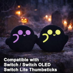 May include: Two black ghost-shaped thumbstick covers with glowing eyes and mouths. One has purple accents, the other yellow. The text at the bottom says "Compatible with Switch / Switch OLED Switch Lite Thumbsticks."
