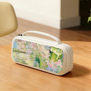May include: A rectangular, off-white pencil case with a handle and zipper closure. The case features a colorful floral and landscape design with pink, blue, and green hues. The case is sitting on a wooden surface.