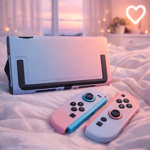 May include: A pastel-coloured gaming console and two controllers. The console has a blue and pink gradient, with a black stand. The controllers are pink and blue, with black buttons. The items are on a white, textured surface.