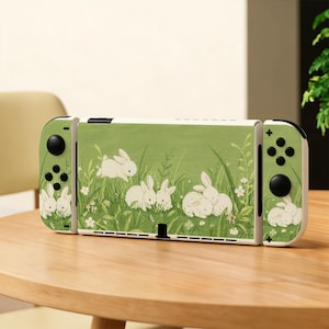 May include: A gaming console featuring a light green skin with a repeating illustration of white rabbits in tall grass. The device includes two detachable controllers on the sides, each with black buttons and joysticks, matching the rabbit pattern.