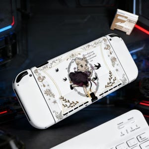 Puede incluir: Consola Nintendo Switch blanca con una piel personalizada con un personaje de zorro de dibujos animados con un vestido morado oscuro. La piel tiene un borde decorativo con motivos florales y de pájaros. El texto de la piel está en fuente de escritura.