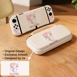 May include: Cream-coloured Nintendo Switch console and carrying case. Both feature a cartoon bunny holding a pink flower, with the text "Good luck." The console has black buttons and a screen. The case has a white zip and handle.