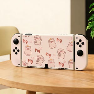 May include: A white gaming console featuring a light pink skin with a repeating pattern of cartoon pigs wearing crowns, some holding drinks, and the word Pig printed in brown text. The console has white detachable controllers on each side.