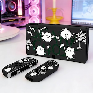 以下が含まれることがあります： ハロウィーンをテーマにした黒いゲーム機ケースとコントローラー。ケースには、白い幽霊とクモの巣のデザインと「SPOOKY SHOW」の文字が施されています。コントローラーには、お揃いの幽霊と足跡のデザインがあります。