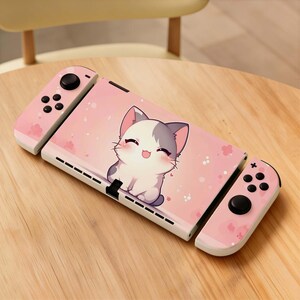 May include: A pink and white Nintendo Switch console with a cute cartoon cat design. The console features pink controllers and a pink cover with a smiling cat illustration and small pink flower accents. The console is on a wooden surface.