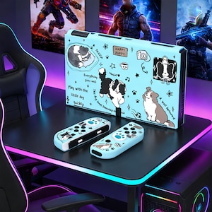 Puede incluir: Una funda de consola de juegos azul claro y dos mandos a juego, decorados con perros de dibujos animados, estrellas y notas musicales. La funda tiene el texto "HAPPY PUPPY" y otras frases. Los mandos y la funda están sobre un escritorio negro.