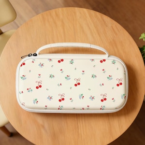 Może przedstawiać: Kremowe etui z uchwytem, ozdobione wzorem czerwonych wiśni i małych gałązek kwiatowych. Etui ma biały zamek błyskawiczny i zaokrąglony prostokątny kształt, odpowiedni dla elektroniki.