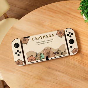 Puede incluir: Una consola Nintendo Switch de color crema con un tema de carpincho. La consola presenta la palabra "CAPYBARA" e ilustraciones de carpinchos. También es visible el texto "Capybara is a little cute guy. Lorem omnis baruku bacok aja bu".