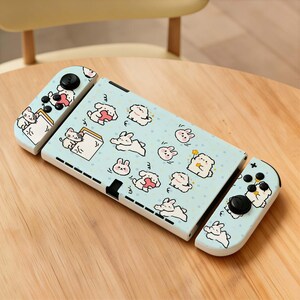 May include: A light blue Nintendo Switch console with a playful design featuring cartoon bunnies and hearts. The controllers and console body are adorned with the cute illustrations. The buttons are black.