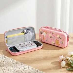 Può includere: Due custodie rosa per console di gioco, una aperta e una chiusa. La custodia aperta mostra la console e scomparti portaoggetti. La custodia chiusa presenta un motivo a gatto e fiori. Su una superficie di legno.