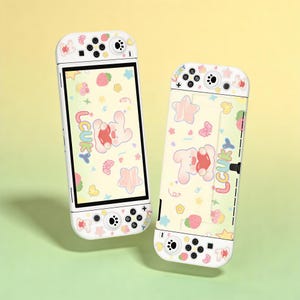 Puede incluir: Dos consolas Nintendo Switch blancas con un diseño pastel. Cada consola presenta un conejito de dibujos animados, estrellas, fresas y la palabra "LCUKY". Los mandos tienen botones negros y un fondo blanco.