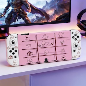Puede incluir: Un mando rosa de Nintendo Switch con un diseño de tres en raya con ilustraciones de gatos. El mando tiene botones blancos y un diseño de huella de pata en el lado izquierdo. El fondo incluye un monitor de ordenador y auriculares.