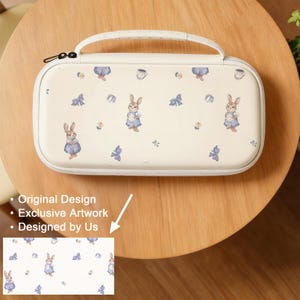 May include: Cream-colored carrying case with a white handle and zipper. The case is decorated with a repeating pattern of bunnies in blue dresses, blue bows, and teacups. The text "Original Design", "Exclusive Artwork", and "Designed by Us" is visible.