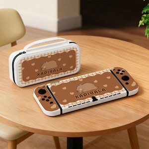 Puede incluir: Un estuche y una funda para consola Nintendo Switch blancos y marrones. El estuche tiene un asa y una cremallera. Ambos artículos presentan un diseño marrón con un oso durmiente, corazones y la palabra "KAPIBALA".