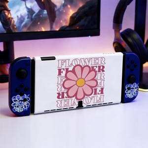 May include: A white and blue Nintendo Switch console with a floral design. The center of the console features a pink flower with the word "FLOWER" in a repeating pattern. The controllers are blue with a flower graphic.