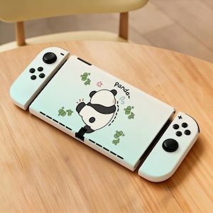 May include: A light blue Nintendo Switch console with a panda design. The console has a white cover with a cartoon panda illustration, green bamboo, and the word "panda" written in a playful font. The controllers are light blue with black buttons.