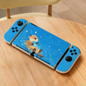 May include: A portable gaming console featuring a blue skin with a brown otter illustration holding a blue fish. The device includes two detachable blue controllers on the sides, each equipped with black joysticks and buttons for gameplay interaction.
