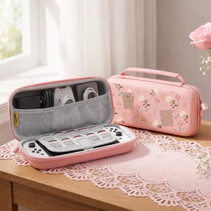 May include: Two pink game console cases. One is open, showing a white console and accessories. The other case is closed, with a bear and floral design. Both cases are on a wooden table with a pink lace runner.