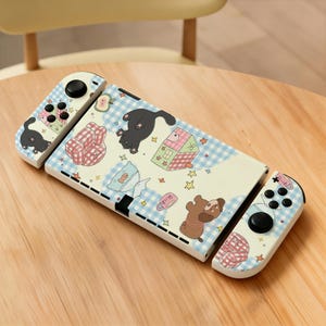 May include: A handheld gaming console featuring a cream-colored skin with a light blue gingham pattern. The design includes illustrations of black cats, brown dogs, pink plaid armchairs, small houses, apples, and yellow stars scattered across the surface.