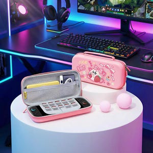 May include: Pink gaming console carrying case with a strawberry and bear design, open to reveal a console and accessories. A second closed case with the text "nice" is also visible. Two pink spheres sit on a white surface.