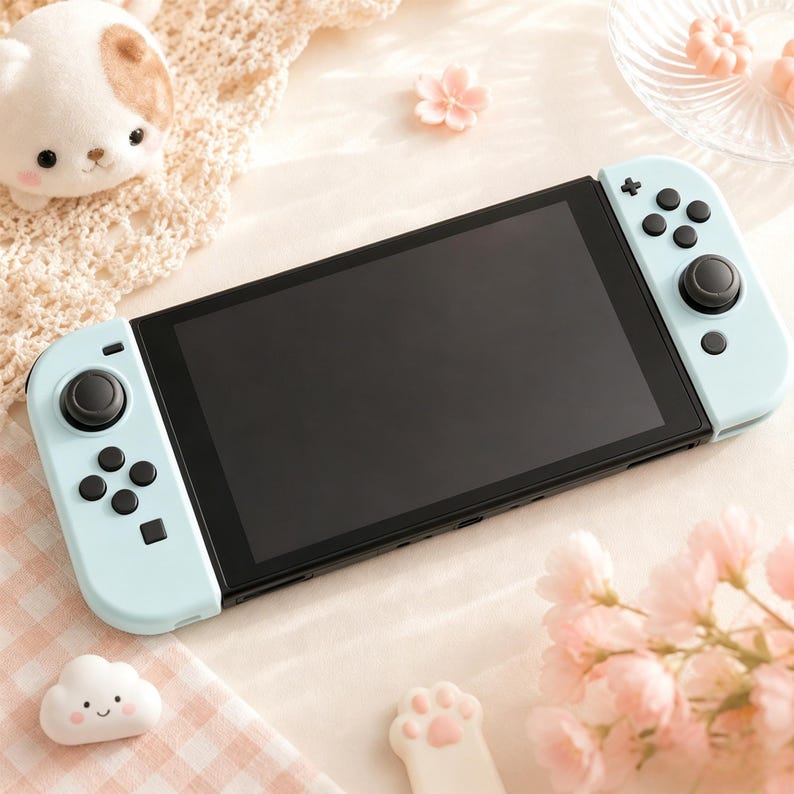 May include: A light blue Nintendo Switch console with black screen and buttons. The console is surrounded by decorative items, including a plush animal, flower-shaped candies, and pink flowers. The background is a soft, light color.