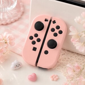 May include: A pair of pink Joy-Con controllers for a gaming console, featuring black buttons and joysticks. The controllers are set against a backdrop of decorative elements, including a cloud-shaped charm, a heart-shaped charm, and floral accents.