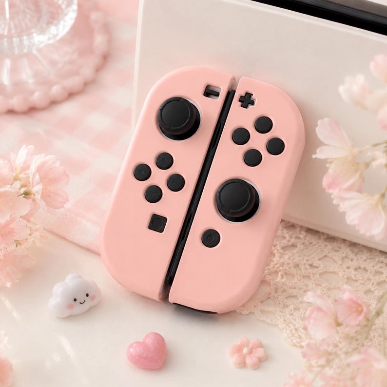 May include: A pair of pink Joy-Con controllers for a gaming console, featuring black buttons and joysticks. The controllers are set against a backdrop of decorative elements, including a cloud-shaped charm, a heart-shaped charm, and floral accents.
