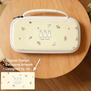 May include: A cream-colored carrying case with a handle, featuring a design of three white rabbits and pink floral accents. The case has a white border and a black zipper. The text "rabbit friends" is below the rabbits.
