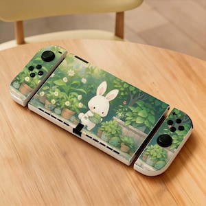 May include: A white Nintendo Switch console with a custom skin featuring a cartoon bunny holding a plant in a garden setting. The controllers and console are decorated with a green and white floral design.