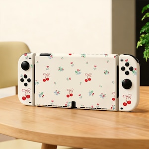 May include: A white Nintendo Switch console with a cream-colored cover featuring a pattern of red cherries and green leaves. The controllers are white with black buttons and the console is on a wooden table.