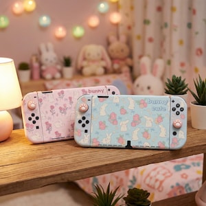 Op de afbeelding: Twee Nintendo Switch-consoles met konijnenontwerpen. Eén is lichtroze met bloemen- en konijnenpatronen, de andere is lichtblauw met konijnen- en aardbeipatronen. De controllers hebben pootvormige knoppen.