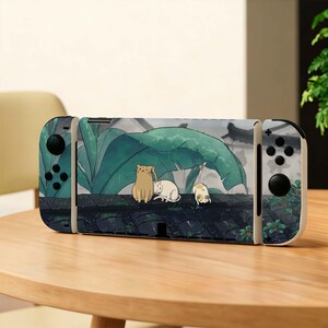 May include: A Nintendo Switch console with a custom skin featuring an illustration of three cats under a large green leaf. The design includes a beige cat, a white cat, and a calico cat. The background has a dark, rainy aesthetic.