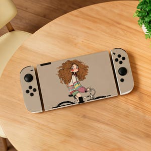 May include: A beige gaming console featuring a decal of a cartoon character with voluminous curly brown hair, wearing a striped sweater and riding a bicycle. The console has two detachable controllers with black buttons and joysticks on each side.