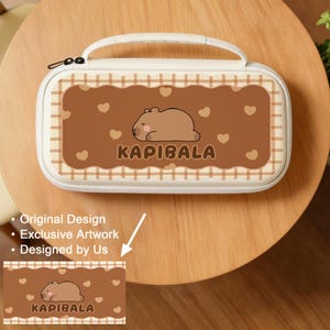 Puede incluir: Una funda de transporte color crema con un diseño rectangular marrón que presenta un capibara de dibujos animados y la palabra "KAPIBALA". La funda tiene un asa blanca y un cierre de cremallera. El diseño incluye formas de corazón.