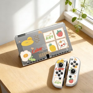 Może przedstawiać: Szare etui i kontrolery do Nintendo Switch z kolorowymi, kreskówkowymi wzorami. Etui przedstawia kaczkę, kwiat i znaczki z truskawką, ananasem i misiem. Kontrolery mają pasujące wzory.
