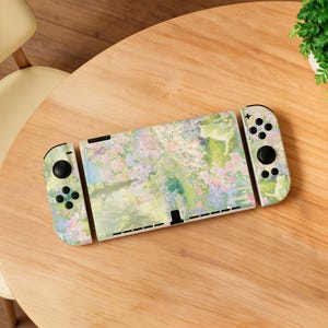 May include: A Nintendo Switch console with a floral design. The console and controllers feature a pastel color palette with pink, green, and yellow hues. The design resembles a watercolor painting of a garden scene.