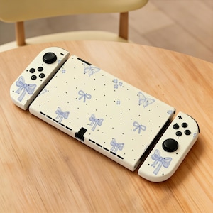 May include: A white Nintendo Switch console with a cream-colored cover and controllers. The cover and controllers are decorated with light blue bows, butterflies, and black polka dots. The console is resting on a wooden surface.
