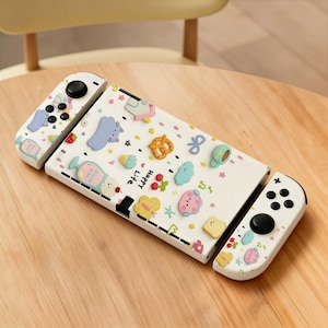 Pode incluir: Uma capa e comandos brancos para Nintendo Switch decorados com desenhos coloridos em relevo de comida e animais de desenho animado. A capa tem a inscrição "Happy Life". Os comandos têm botões e joysticks pretos.