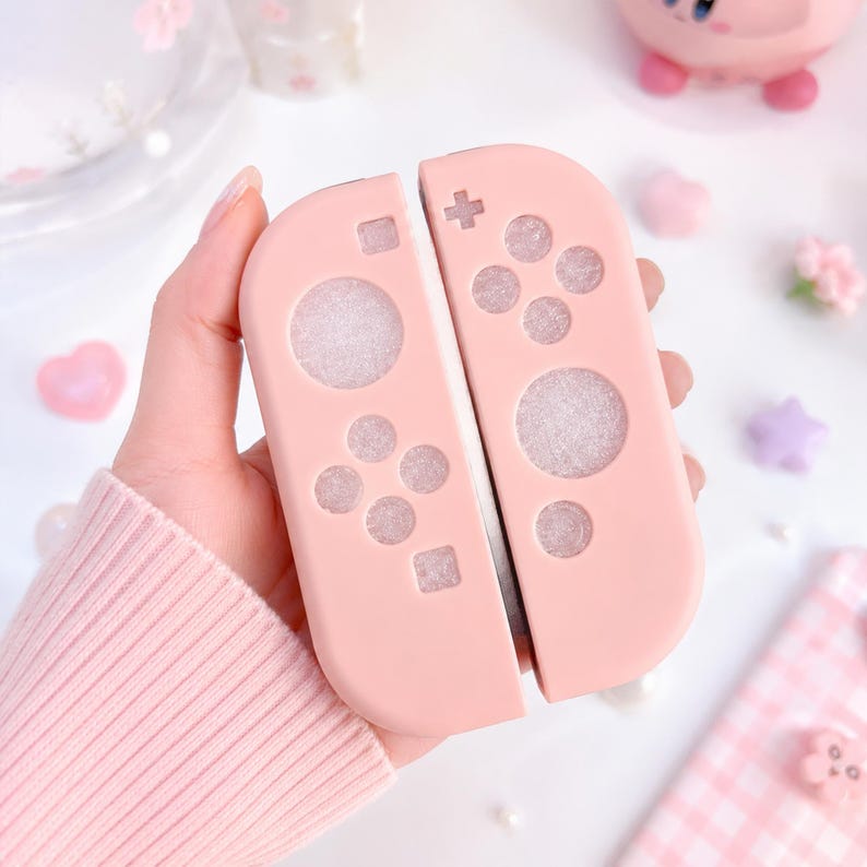 May include: A pair of light pink Nintendo Switch Joy-Con controllers. The controllers have a matte finish and feature the standard button layout, including directional pads and action buttons. The background includes decorative elements like hearts and stars.