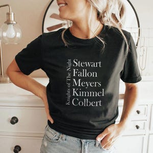 Puede incluir: Camiseta negra de cuello redondo con el texto "Knights of The Night" y los nombres Stewart, Fallon, Meyers, Kimmel y Colbert en blanco. Una camiseta de manga corta.