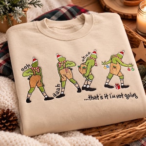 May include: A beige crewneck sweatshirt featuring embroidered green characters wearing Santa hats and tan overalls. The design shows four sequential poses with the text "ooh", "aaah", "mhmm", and "...that's it i'm not going" stitched in black thread.