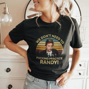 May include: Black t-shirt featuring a retro design with the text "WE DON'T NEED TO FUCKING PRACTICE RANDY!". The design includes a portrait of a man wearing a baseball cap, set against a sunset background.