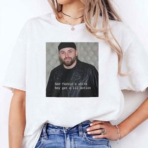 May include: White t-shirt with a square image of a man in a black hat and jacket. The image includes the text "God forbid a white boy get a lil motion". The shirt is a casual style.